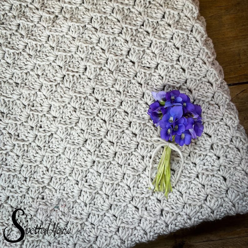 Puede incluir: Primer plano de una manta de ganchillo de color beige claro con un peque&ntilde;o ramo de flores moradas en la parte superior. La manta tiene un patr&oacute;n texturizado en forma de diamante.