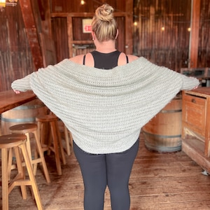 May include: A woman wearing a light gray knitted cardigan with a loose, draped fit. The cardigan is worn over a black tank top and black leggings.