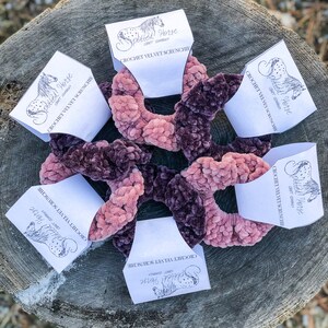 May include: Five crocheted velvet scrunchies in shades of pink and purple, displayed on a wooden surface. Each scrunchie is packaged with a white card featuring the logo "Spotted Horse Craft Company" and the text "Crochet Velvet Scrunchie".