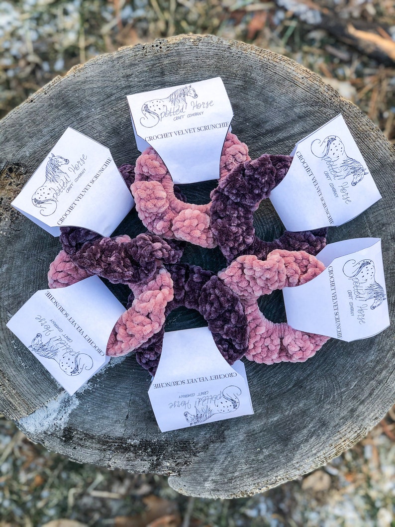 May include: Five crocheted velvet scrunchies in shades of pink and purple, displayed on a wooden surface. Each scrunchie is packaged with a white card featuring the logo "Spotted Horse Craft Company" and the text "Crochet Velvet Scrunchie".