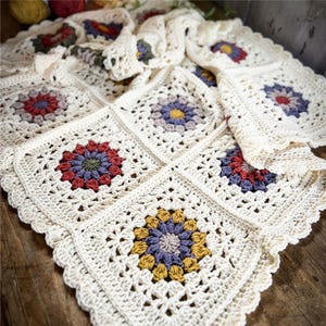 Puede incluir: Manta de ganchillo hecha a mano, de color crema, con diseños florales coloridos. La manta está hecha de motivos cuadrados individuales unidos. Las flores son en tonos de rojo, morado, amarillo y verde. Los bordes tienen un borde festoneado.