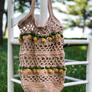 Puede incluir: Una bolsa de mano de crochet beige con pompones amarillos y verdes. La bolsa est&aacute; colgada de una silla blanca.