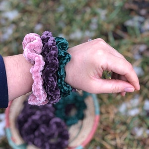 May include: Three velvet scrunchies in pink, purple, and green colors. The scrunchies are stacked on a person's wrist.