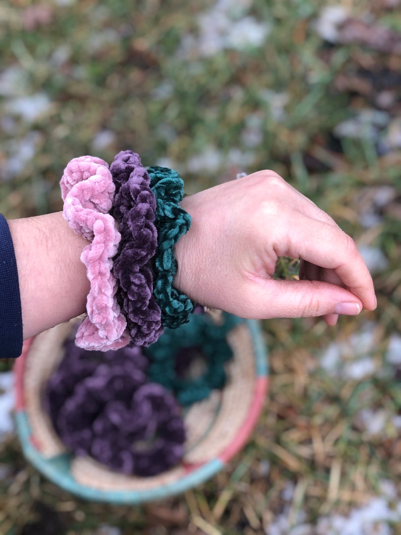 May include: Three velvet scrunchies in pink, purple, and green colors. The scrunchies are stacked on a person's wrist.