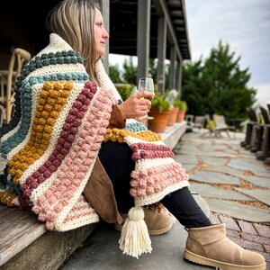 以下が含まれることがあります： 女性が、大きなタッセルが付いたカラフルな編み物の毛布にくるまって、ポーチに座っています。彼女は白ワインのグラスを持って、景色を眺めています。