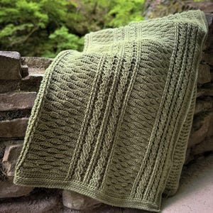 May include: A sage green crochet blanket featuring a textured pattern of vertical columns. The design incorporates alternating sections of raised cable-like stitches and intricate, repeating wave-like motifs throughout the entire fabric surface.