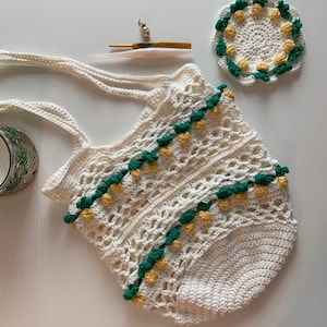 Puede incluir: Una bolsa de mano de crochet blanca con detalles verdes y amarillos. La bolsa tiene una parte superior ancha y abierta y dos asas largas. La bolsa est&aacute; decorada con un patr&oacute;n de peque&ntilde;as flores de crochet.