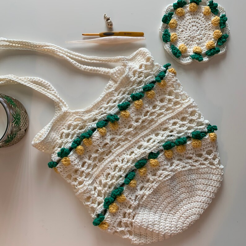 Puede incluir: Una bolsa de mano de crochet blanca con detalles verdes y amarillos. La bolsa tiene una parte superior ancha y abierta y dos asas largas. La bolsa est&aacute; decorada con un patr&oacute;n de peque&ntilde;as flores de crochet.