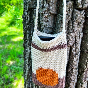 May include: A crocheted phone pouch with a brown, white, and orange striped design. The pouch is hanging from a tree trunk.
