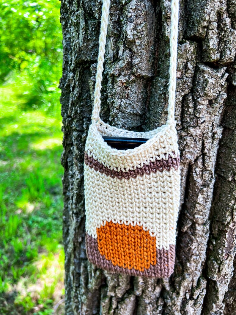 May include: A crocheted phone pouch with a brown, white, and orange striped design. The pouch is hanging from a tree trunk.