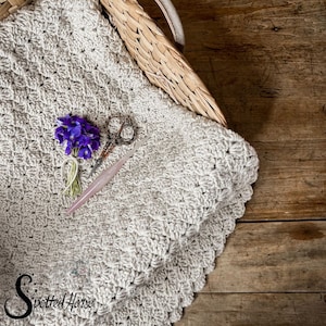 Puede incluir: Una manta de ganchillo beige con un borde festoneado, un par de tijeras antiguas y un ramo de flores moradas. La manta est&aacute; en una cesta de mimbre sobre una superficie de madera.