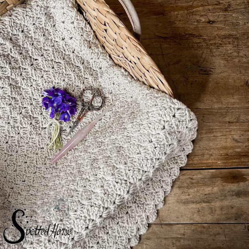 Puede incluir: Una manta de ganchillo beige con un borde festoneado, un par de tijeras antiguas y un ramo de flores moradas. La manta est&aacute; en una cesta de mimbre sobre una superficie de madera.