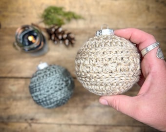 May include: Two crocheted Christmas baubles, one is beige and the other is grey. The beige bauble is being held in a hand.