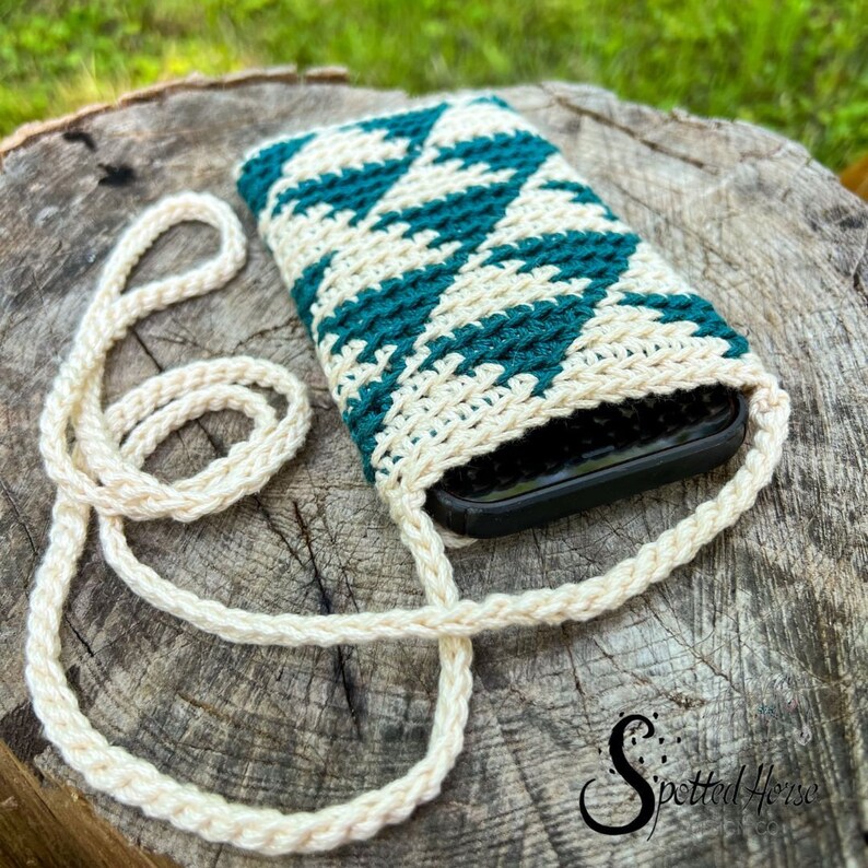 Puede incluir: Una funda de tel&eacute;fono de ganchillo con un patr&oacute;n geom&eacute;trico en color turquesa y crema. La funda tiene una larga correa de color crema para llevarla.