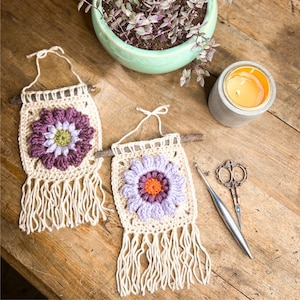 May include: Two cream-coloured crochet wall hangings with flower designs in purple, lavender, and orange. The wall hangings have fringe and are attached to a wooden dowel. A lit candle, scissors, and a crochet hook are also visible.