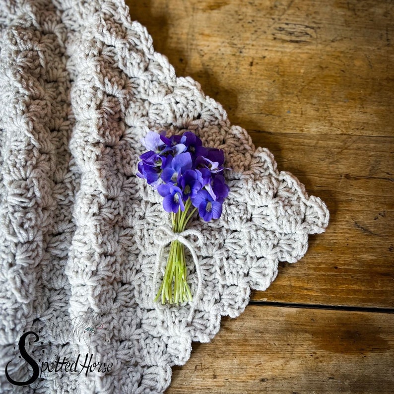 Puede incluir: Un primer plano de una manta de ganchillo con un borde festoneado, que presenta un peque&ntilde;o ramo de flores moradas atadas con una cuerda. La manta es de color gris claro y las flores son de un morado vibrante.