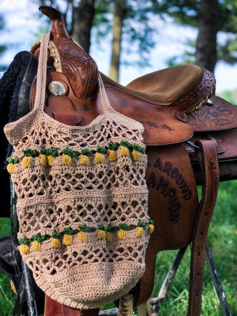 Puede incluir: Una bolsa de mano de crochet con un dise&ntilde;o de pi&ntilde;a cuelga de una silla de montar de cuero marr&oacute;n. La bolsa es beige con pi&ntilde;as amarillas y verdes.