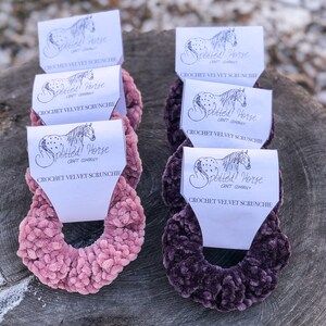 May include: Two crocheted velvet scrunchies, one in pink and one in purple, with a white label that says "Spotted Horse Craft Company" and "Crochet Velvet Scrunchie".