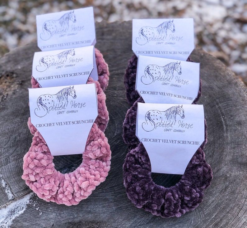 May include: Two crocheted velvet scrunchies, one in pink and one in purple, with a white label that says "Spotted Horse Craft Company" and "Crochet Velvet Scrunchie".