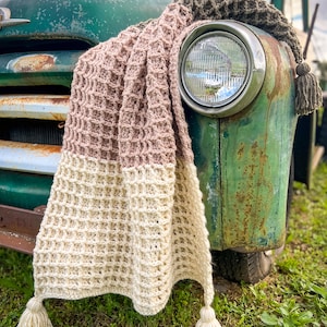 Op de afbeelding: Een gehaakt deken in beige, roze en bruine tinten met kwastjes die aan de onderste hoeken hangen. Het deken is over de voorkant van een vintage groene vrachtwagen gelegd.