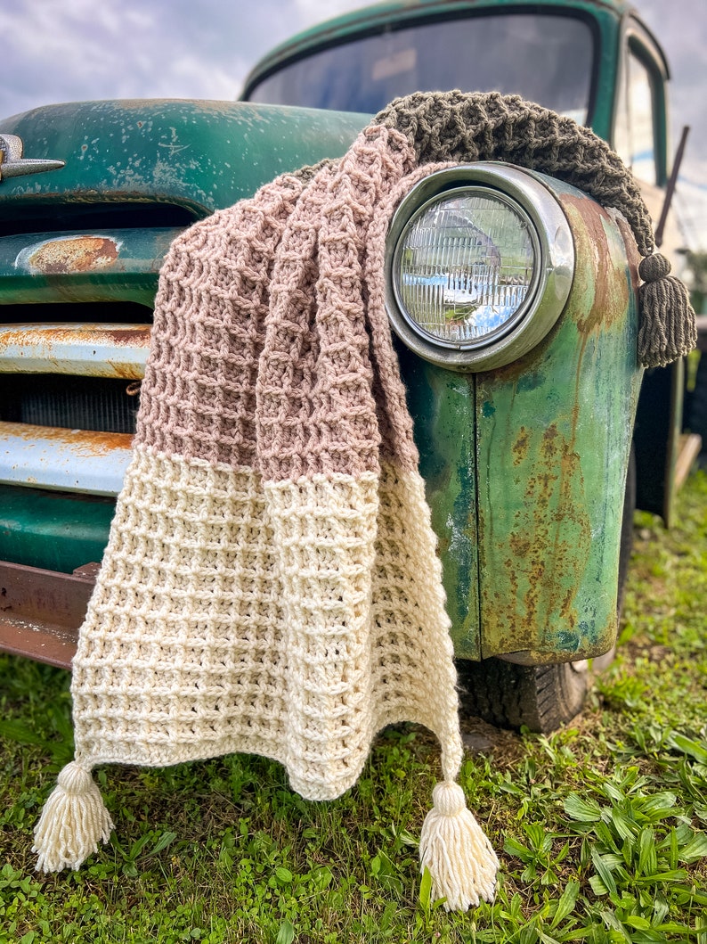 Op de afbeelding: Een gehaakt deken in beige, roze en bruine tinten met kwastjes die aan de onderste hoeken hangen. Het deken is over de voorkant van een vintage groene vrachtwagen gelegd.