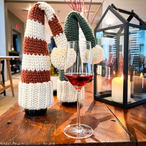 Puede incluir: Dos botellas de vino con tapones de botella de punto en rayas rojas y blancas y verdes y blancas. Una copa de vino tinto se encuentra sobre una mesa de madera con una linterna negra y una vela blanca.