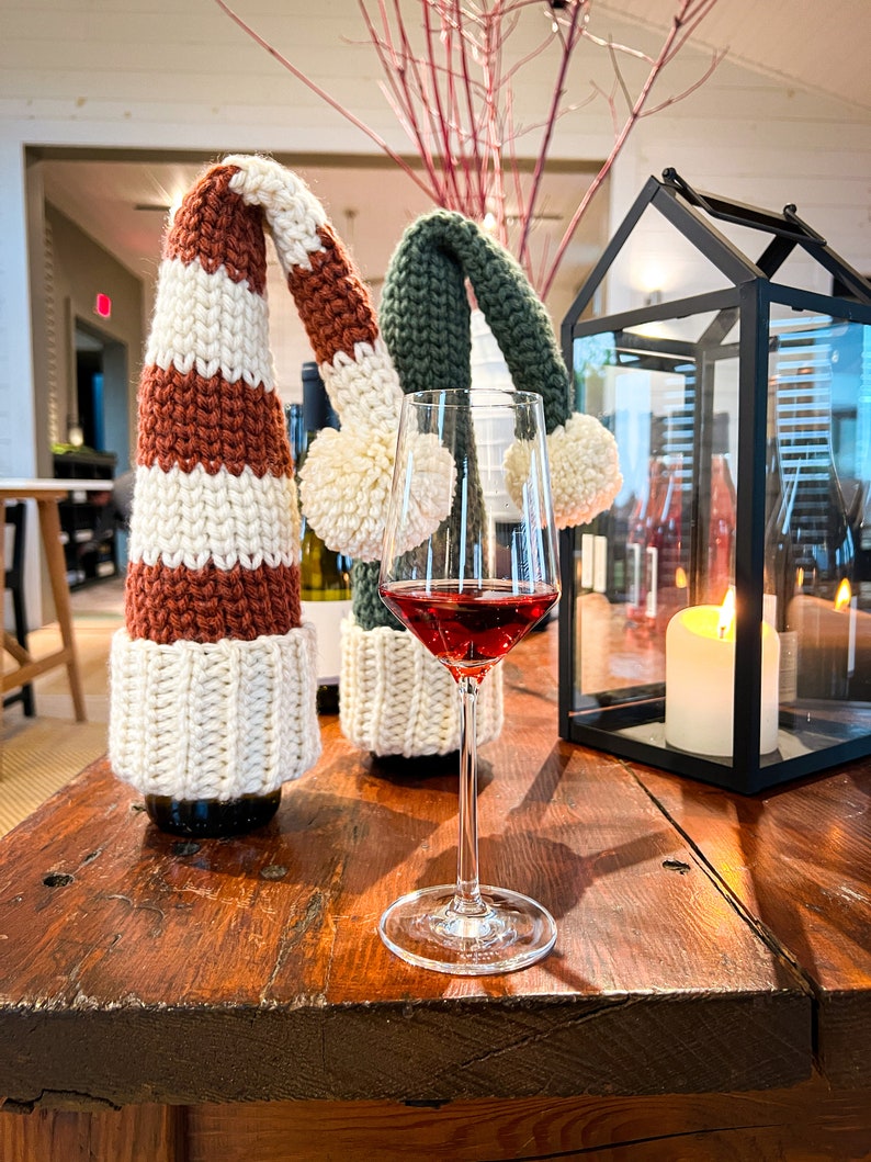 Puede incluir: Dos botellas de vino con tapones de botella de punto en rayas rojas y blancas y verdes y blancas. Una copa de vino tinto se encuentra sobre una mesa de madera con una linterna negra y una vela blanca.