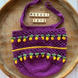 Puede incluir: Bolso de mercado de crochet morado con detalles de pi&ntilde;a amarillos y verdes. El bolso tiene las palabras "MARKET TOTE" escritas en &eacute;l.
