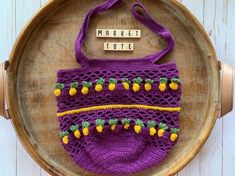 Puede incluir: Bolso de mercado de crochet morado con detalles de pi&ntilde;a amarillos y verdes. El bolso tiene las palabras "MARKET TOTE" escritas en &eacute;l.