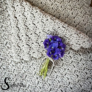 Puede incluir: Un primer plano de una manta de ganchillo de color beige claro con un peque&ntilde;o ramo de flores moradas en la parte superior. La manta tiene un patr&oacute;n texturizado y est&aacute; hecha con un hilo grueso.