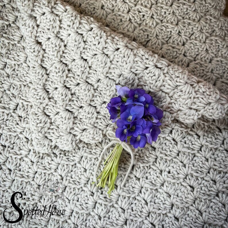 Puede incluir: Un primer plano de una manta de ganchillo de color beige claro con un peque&ntilde;o ramo de flores moradas en la parte superior. La manta tiene un patr&oacute;n texturizado y est&aacute; hecha con un hilo grueso.