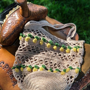 Puede incluir: Una bolsa de mano de crochet con fondo beige y detalles de pi&ntilde;a amarillos y verdes. La bolsa est&aacute; sentada sobre una silla de montar de cuero marr&oacute;n.