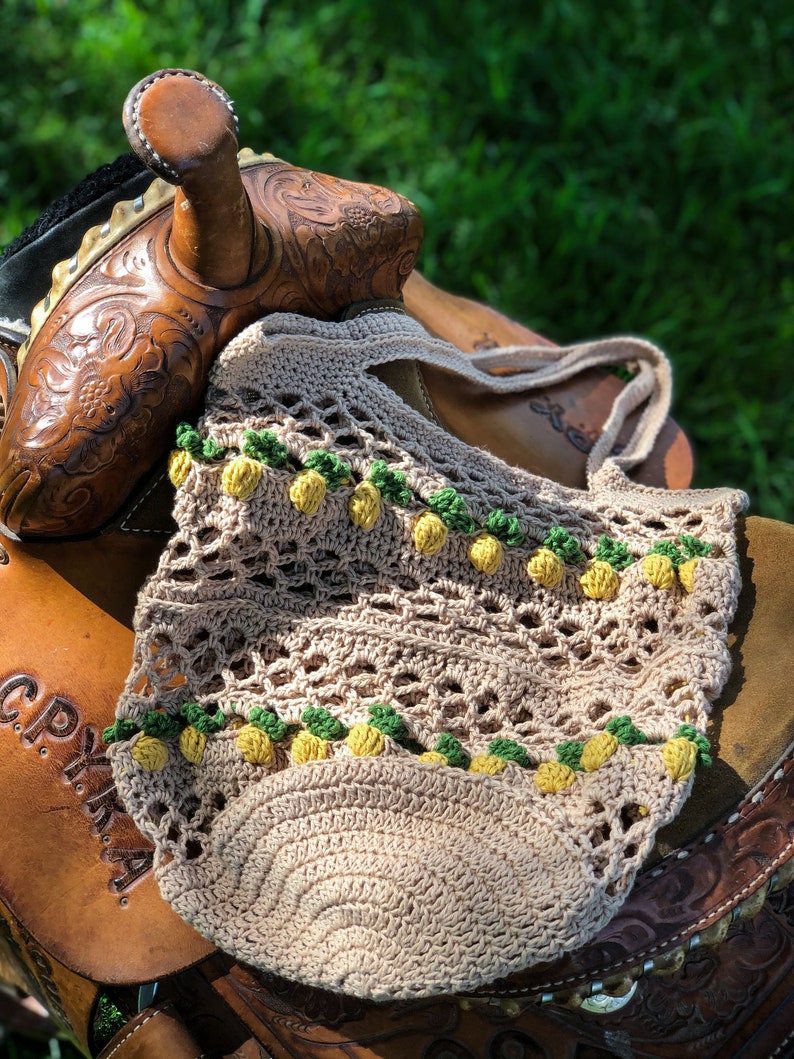 Puede incluir: Una bolsa de mano de crochet con fondo beige y detalles de pi&ntilde;a amarillos y verdes. La bolsa est&aacute; sentada sobre una silla de montar de cuero marr&oacute;n.
