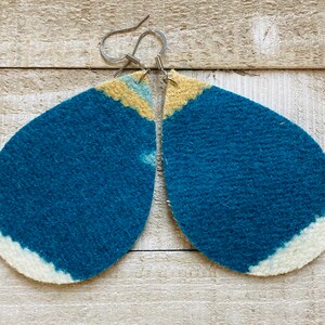 May include: A pair of teal blue felt earrings with a white and yellow ombre design. The earrings are teardrop shaped and have silver hooks.
