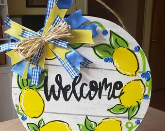 May include: A round wooden door hanger with a whitewashed background and a painted lemon wreath. The wreath features yellow lemons, green leaves, and blue berries. The word "Welcome" is written in black cursive script. A yellow and blue bow with a straw center is attached to the top.