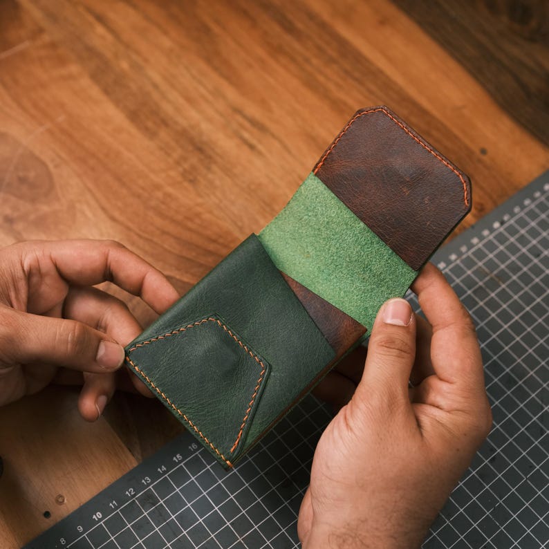 K&ouml;nnte beinhalten: Handgefertigte Ledergeldb&ouml;rse in Gr&uuml;n- und Braunt&ouml;nen mit orangefarbenen N&auml;hten. Die Geldb&ouml;rse ist ge&ouml;ffnet und zeigt einen gr&uuml;nen Innenraum und braune Akzente. Das Design hat eine geometrische Klappe und einen minimalistischen Stil f&uuml;r Karten und Bargeld.