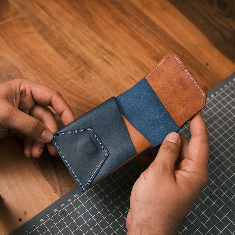 K&ouml;nnte beinhalten: Handgefertigte Ledergeldb&ouml;rse in Marineblau, Hellbraun und Blau. Die Geldb&ouml;rse hat ein geometrisches Design mit sichtbaren N&auml;hten. Die Geldb&ouml;rse wird offen gehalten und zeigt ihr Inneres und ihre Konstruktion.