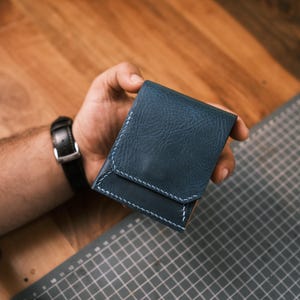 K&ouml;nnte beinhalten: Ein marineblaues Lederportemonnaie mit wei&szlig;en N&auml;hten. Das Portemonnaie wird in einer Hand gehalten und zeigt sein kompaktes Design und seine Handwerkskunst. Es hat einen Klappenverschluss und ein minimalistisches Aussehen.