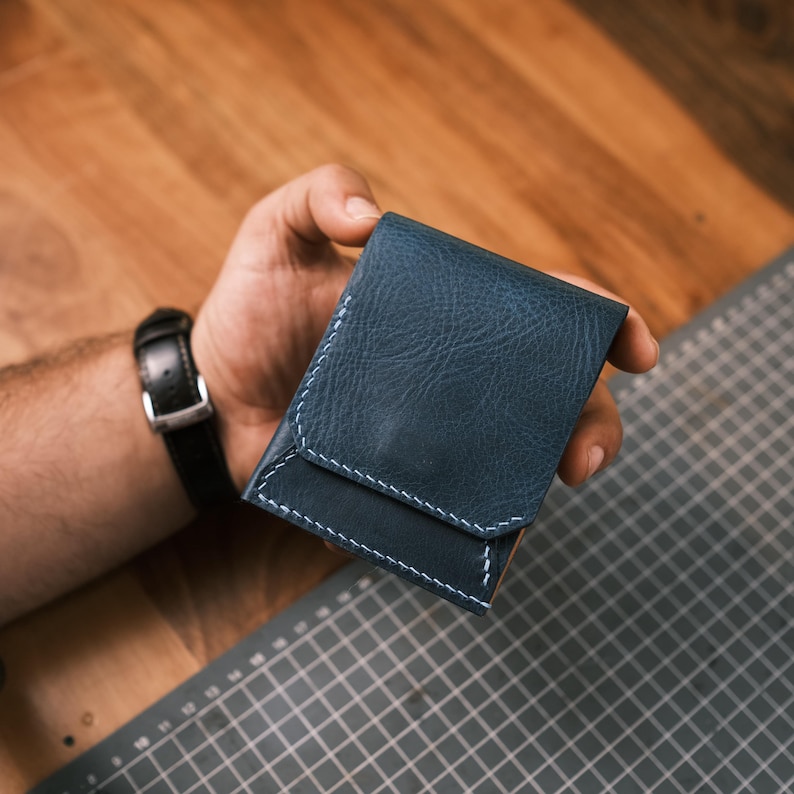 K&ouml;nnte beinhalten: Ein marineblaues Lederportemonnaie mit wei&szlig;en N&auml;hten. Das Portemonnaie wird in einer Hand gehalten und zeigt sein kompaktes Design und seine Handwerkskunst. Es hat einen Klappenverschluss und ein minimalistisches Aussehen.