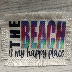 May include: A white fabric patch with frayed edges featuring the text "THE BEACH is my happy place" in black lettering. The word "BEACH" is printed in rainbow colors.