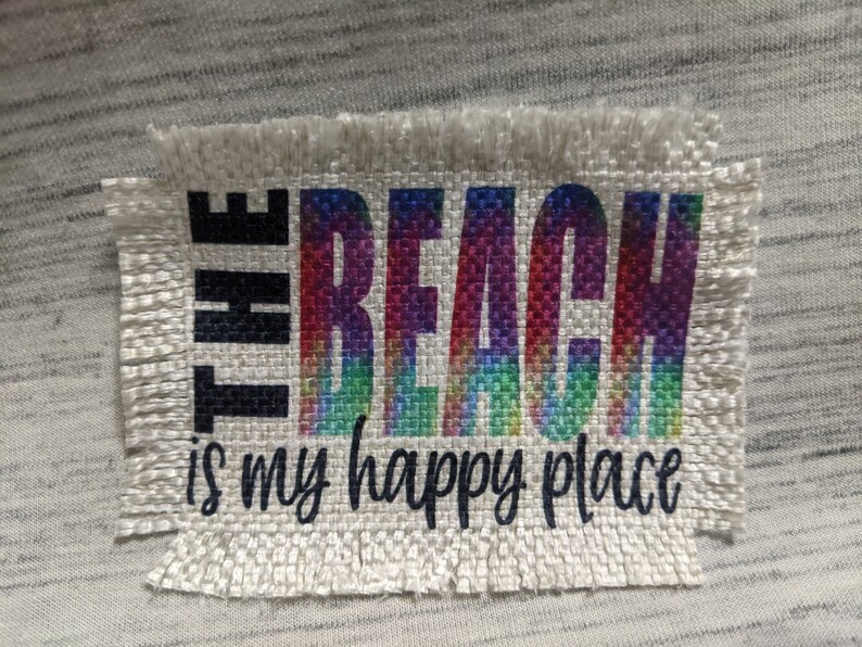 May include: A white fabric patch with frayed edges featuring the text "THE BEACH is my happy place" in black lettering. The word "BEACH" is printed in rainbow colors.