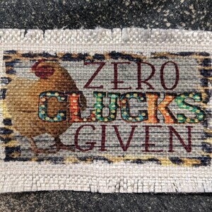 Może przedstawiać: Beżowy kuchenny ręcznik z tkaniny z ilustracją brązowego i białego kurczaka i tekstem "ZERO CLUCKS GIVEN" w czerwonych i czarnych literach.