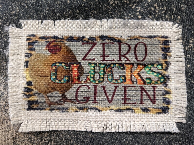 Może przedstawiać: Beżowy kuchenny ręcznik z tkaniny z ilustracją brązowego i białego kurczaka i tekstem "ZERO CLUCKS GIVEN" w czerwonych i czarnych literach.