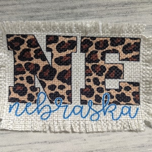 May include: A burlap patch with a frayed edge featuring the letters "NE" in a leopard print design. The letters are outlined in black and the word "Nebraska" is written in blue cursive below the letters.