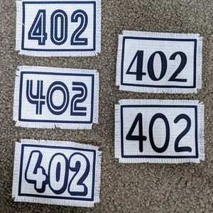May include: Five white fabric patches with black numbers "402" printed on them. The patches have a frayed edge and are arranged in a grid pattern.