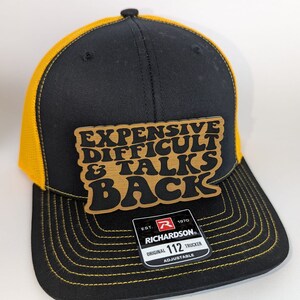 May include: Black and gold trucker hat with a brown patch that says "Expensive Difficult & Talks Back". The hat has a black mesh back and a yellow brim. The hat is adjustable and has a Richardson 112 label.