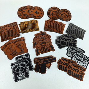 May include: A collection of leather patches with various designs and sayings. Some patches feature American flags, guns, and the words "Try that in a small town". Other patches feature the words "Good girls carry guns" and "Messy buns and guns".