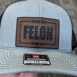 May include: A blue and black trucker hat with a brown leather patch that says "I'm voting for the FELON TRUMP 2024". The hat has a black mesh back and an adjustable strap with the Richardson logo and "112 TRUCKER" text.