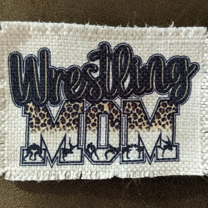 May include: A rectangular patch with the words "Wrestling MOM" in a stylized font. The word "Wrestling" is in dark blue, and "MOM" has a leopard print pattern. Small wrestling figures are at the bottom of the patch.