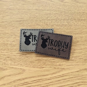 May include: Two brown leather patches with a stitched border. One patch has a silhouette of a deer head with antlers and the text "Trophy Wife". The other patch has a silhouette of a deer head with antlers and the text "Trophy Wife".